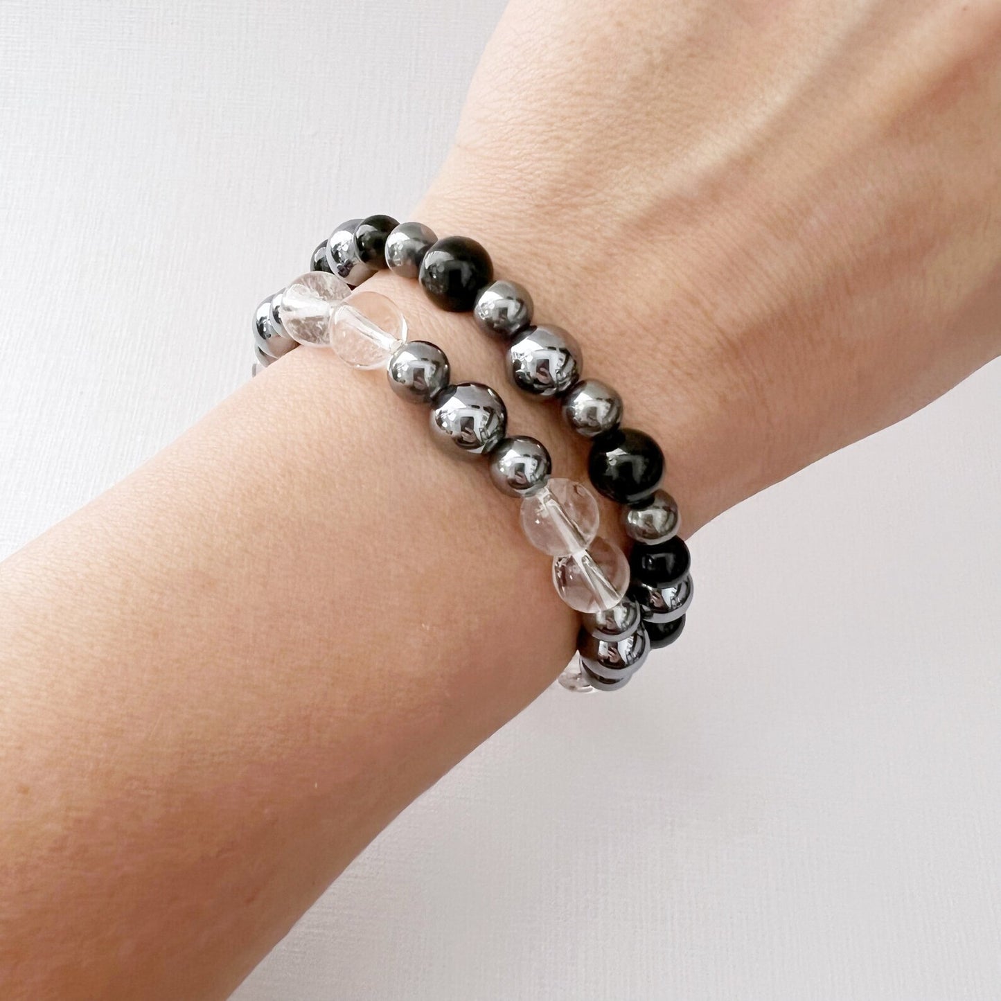 Two **8mm Terahertz Beaded Bracelets** are displayed on a white surface. The bracelet on the left features alternating Clear Quartz and silver beads, while the bracelet on the right consists of alternating Black Tourmaline and silver beads. Both bracelets have a minimalist and elegant design.