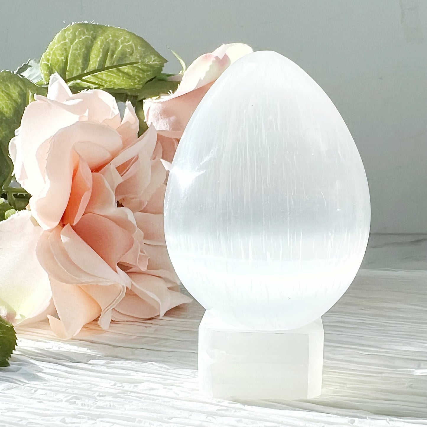 A smooth, egg-shaped 2.5" Selenite Egg stands upright on a matching rectangular base. The setup is placed on a white surface with soft pink roses and green leaves in the background, creating a serene and elegant display, perfect as a meditation accessory for spiritual growth.