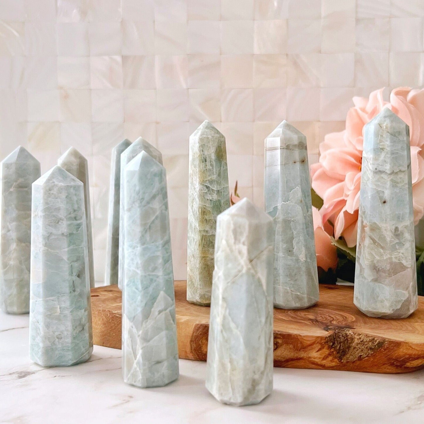 A collection of polished crystal towers, including a stunning 3-4" Aquamarine Standing Point, are displayed on a wooden slab. Primarily light blue and green, they promote emotional balance. Behind them, blush pink flowers and a white tiled wall create a tranquil and aesthetically pleasing backdrop.