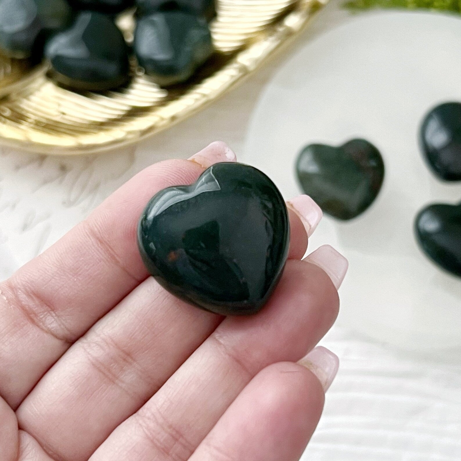 A set of small, polished 1" Mini Bloodstone Hearts displayed on a golden dish and a white circular platform. The background includes green foliage and a piece of paper with cursive text. These stones offer grounding and emotional support, with one heart-shaped stone placed beside the platform.