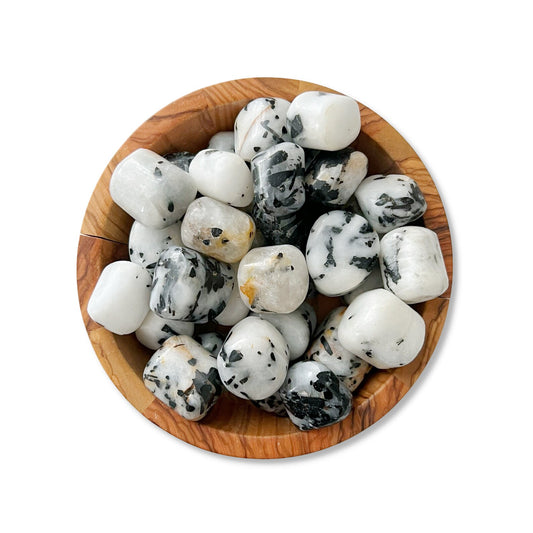 A wooden bowl filled with Tourmalated Quartz Tumbled Stones, primarily white with black speckles, sits against a plain white background. The stones have a smooth, glossy finish, and the natural grain of the wooden bowl contrasts with the stones' patterns, symbolizing spiritual growth and protection from EMF pollution.