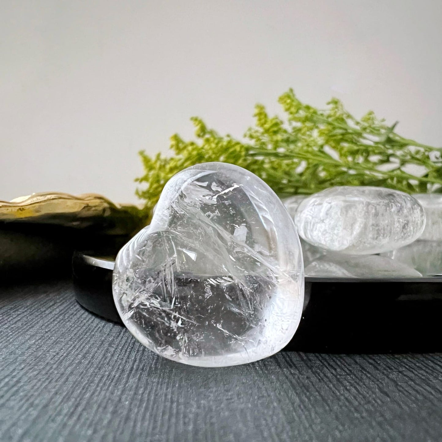 Two 1" Mini Clear Quartz Hearts rest on a wooden surface, surrounded by green foliage and small buds. The hearts, reminiscent of clear quartz treasures, showing clear and cracked textures. The overall composition is earthy and natural.