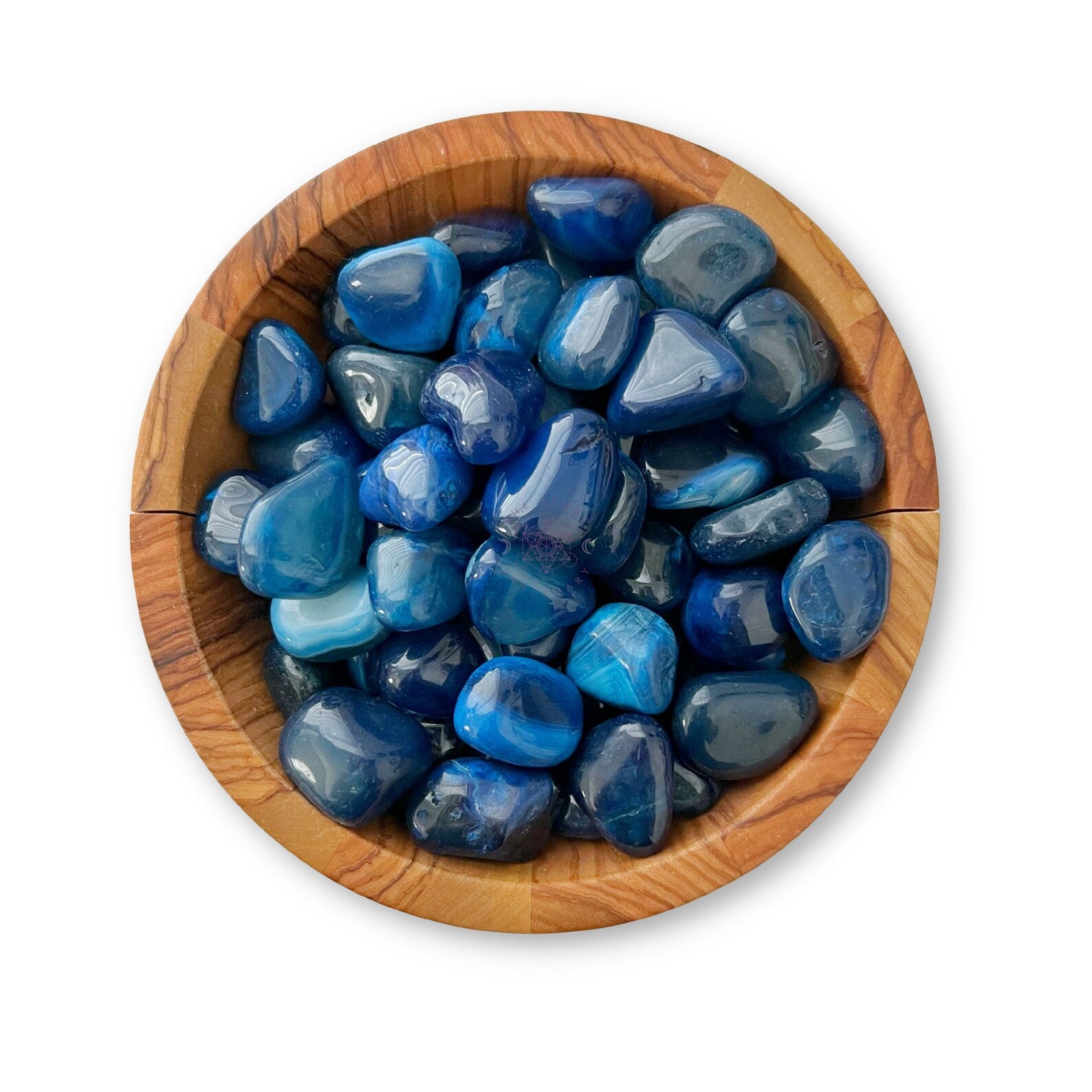 A wooden bowl filled with Blue Agate Tumbled Stones of various shades and shapes. The stones, known for their throat healer properties, exhibit a glossy finish. The bowl has distinct grain patterns, giving it a rustic yet elegant appearance. The plain white background makes the colors stand out vividly.