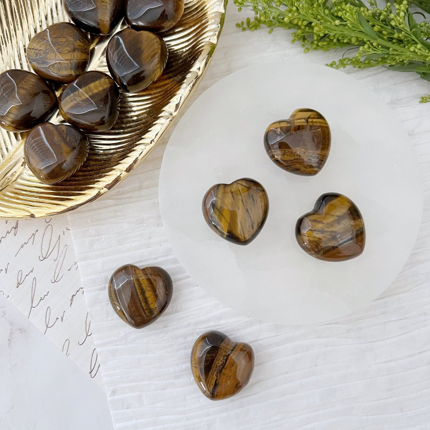 Displayed on white textured surfaces with green foliage in the background, a collection of 1" Mini Tiger Eye Hearts are showcased elegantly. Some of these polished, heart-shaped stones rest on a round clear plate, while others are placed on a gold leaf-shaped tray. Handwritten notes and personalized jewelry ideas can be seen partially in the frame.