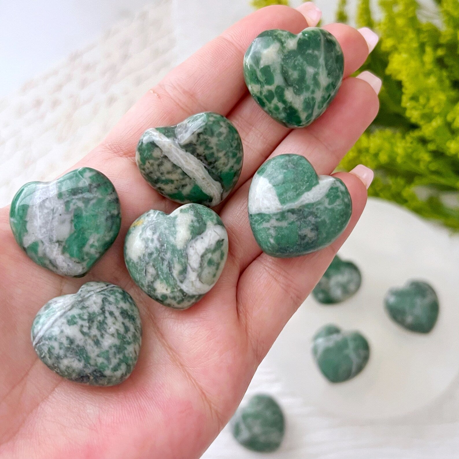 A display of green and white marbled heart-shaped stones arranged on different surfaces. Four 1" Mini Rich Jade Hearts are placed on a white round platform, one lies beside it, and several more polished gemstone hearts sit on a golden dish. Green foliage adds a decorative touch in the background.