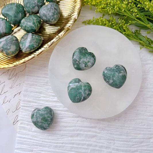 A display of green and white marbled heart-shaped stones arranged on different surfaces. Four 1" Mini Rich Jade Hearts are placed on a white round platform, one lies beside it, and several more polished gemstone hearts sit on a golden dish. Green foliage adds a decorative touch in the background.