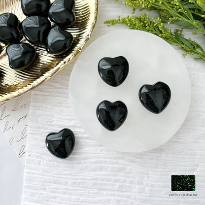 A round white plate on a white textured surface holds four polished, black heart-shaped stones. Nearby, a gold tray filled with additional heart stones and green foliage is visible. A patch of black stone with hints of green, known for its Heart Chakra benefits and emotional healing properties, is labeled "1" Mini Green Goldstone Heart.