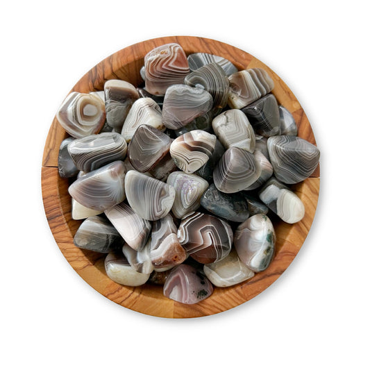 A wooden bowl filled with numerous polished, multicolored Botswana Agate Tumbled Stones showcases the beauty of Botswana Agate. The stones display a variety of intricate patterns and shades of grey, brown, white, and tan, highlighting their natural banding and protective qualities. The bowl sits on a plain white background.