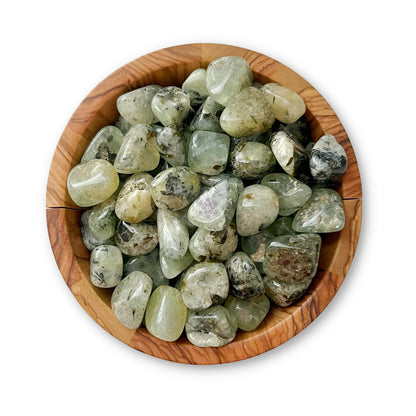 A wooden bowl filled with polished, light green Prehnite Tumbled Stones featuring varying patterns and shades, some with darker inclusions. The tumbled stones are set against a white background.