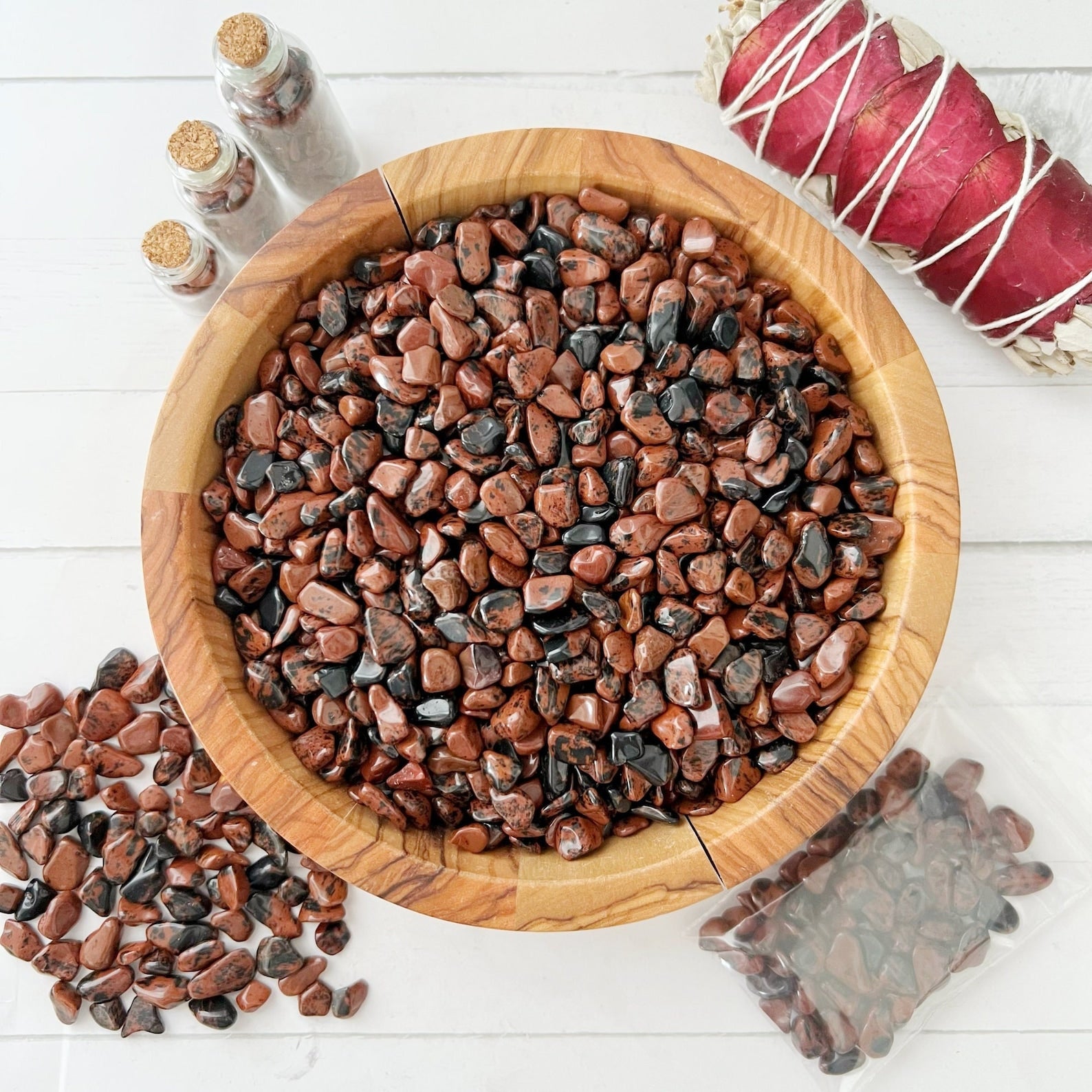 A wooden bowl filled with polished red and black Mahogany Obsidian Crystal Chips sits on a white surface. Nearby, an open bag of similar stones, small glass bottles with cork stoppers, and a bundle wrapped in red and white string are placed neatly around the Mahogany Obsidian Crystal Chips, perfect for crafting resin jewelry.