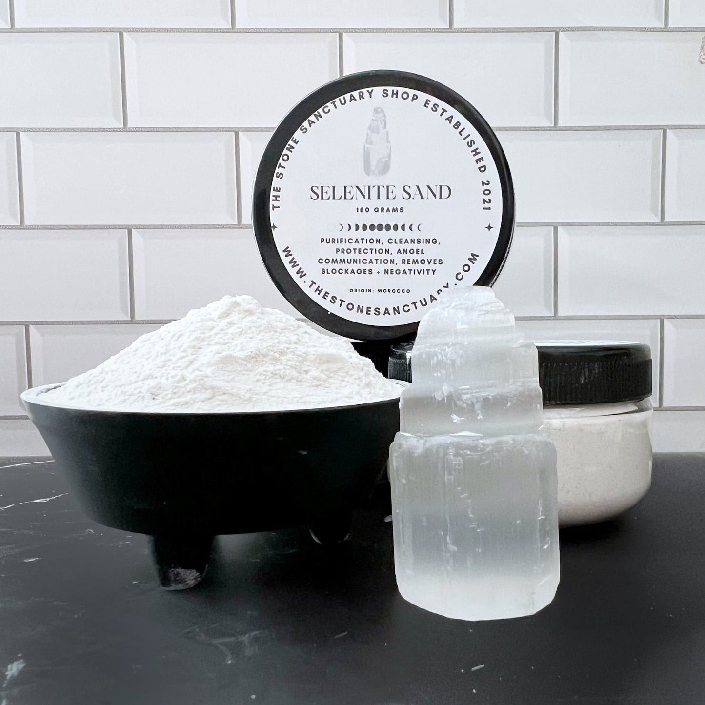 A black bowl filled with Selenite Powder Sand, a cylindrical container of selenite powder, and a stacked selenite crystal gemstone are set against a white tiled backdrop. A circular label in the background reads “Selenite Sand” and provides details about its uses and healing properties.