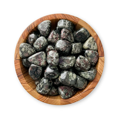 A wooden bowl filled with polished, smooth stones featuring a mix of dark and light streaks and patches in various shades of gray, black, and maroon. This exquisite crystal collection includes Eudialyte Tumbled Stones. The bowl sits against a white background.