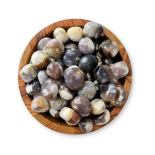 A wooden bowl filled with Purple Chalcedony Tumbled Stones, including purple chalcedony, stands against a plain white background. The stones, varying in hues of purple, gray, beige, and white, have smooth surfaces and a glossy finish—perfect for meditation or psychic protection.