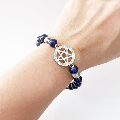 An 8mm Lapis Lazuli & Clear Quartz Beaded Bracelet with Pentacle Charm rests on a wooden surface. In the background, there’s a branch with soft, fuzzy buds and a textured green cloth, creating a rustic and natural setting that highlights this energy bracelet.