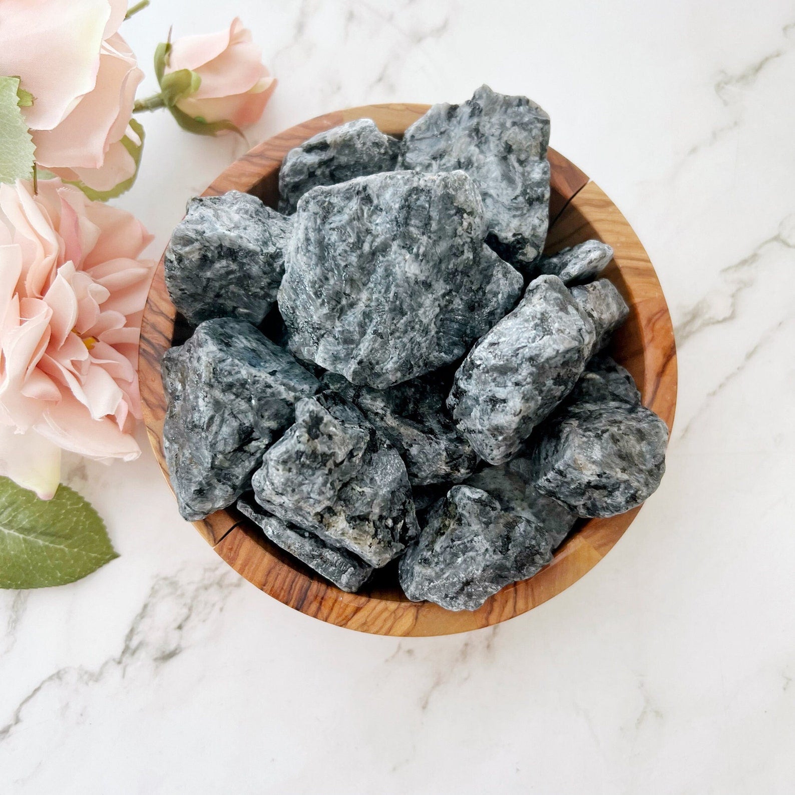 A wooden bowl filled with rough, dark gray Raw Larvikite Stones sits on a white marble surface. Next to the bowl, light pink roses and green leaves add a touch of color and contrast to the scene, perfect for any crystal collection aiming for inner transformation.