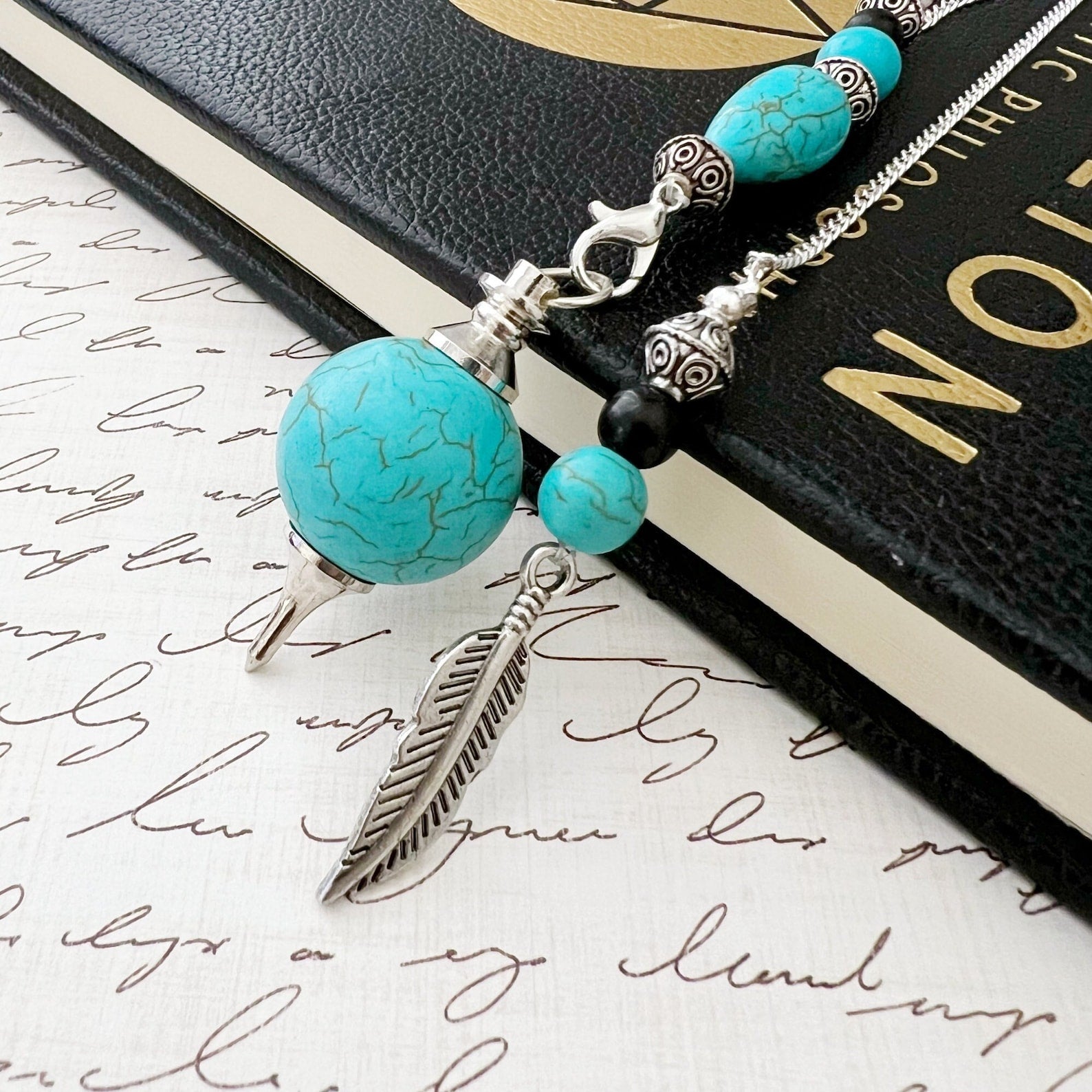 A Turquoise Howlite Seferoth Pendulum with turquoise howlite and black agate beads, silver accents, and a feather charm rests on a journal with cursive writing. The journal's cover is partially visible, featuring elegant black and gold detailing, perfect for those interested in energy balancing and divination.