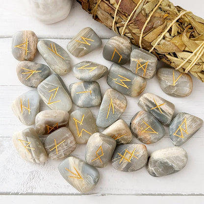 A collection of Black Moonstone Elder Futhark Runes with gold-etched symbols is scattered on a white surface. In the background, there is a white skull decoration, a bundle of sage, and a small potted succulent with pink leaves.