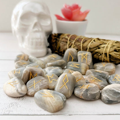 A collection of Black Moonstone Elder Futhark Runes with gold-etched symbols is scattered on a white surface. In the background, there is a white skull decoration, a bundle of sage, and a small potted succulent with pink leaves.