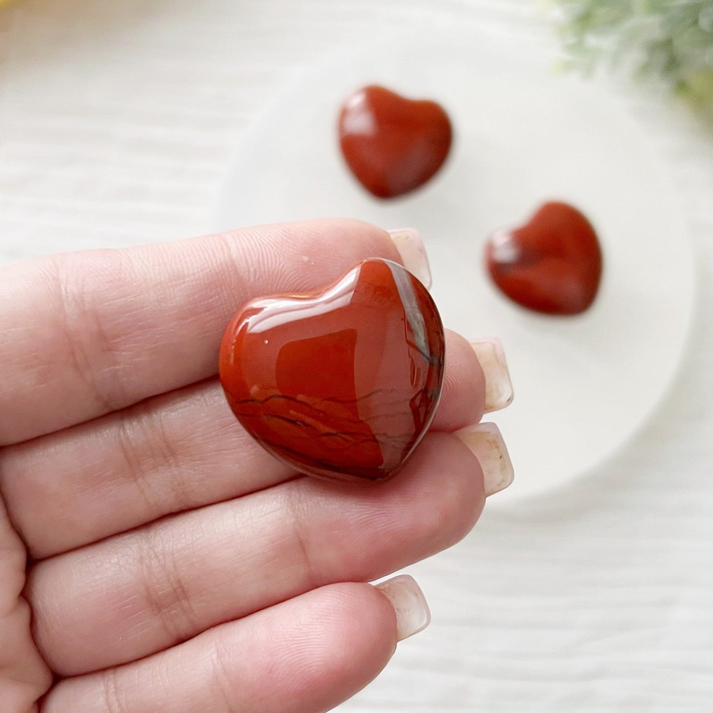 A decorative setup featuring red heart-shaped confections and 1" Mini Red Jasper Hearts. Several hearts are placed on a round white marble platter, with more in a gold dish nearby. The arrangement is on a white crumpled sheet, accented with greenery and a handwritten script underneath.