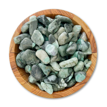 A wooden bowl filled with various tumbled green gemstones, including Emerald Tumbled Stones. The Emerald Tumbled Stones vary in shades of green, some appearing dark with specks of lighter colors and others lighter and more translucent. The bowl is placed against a white background, highlighting their metaphysical properties.