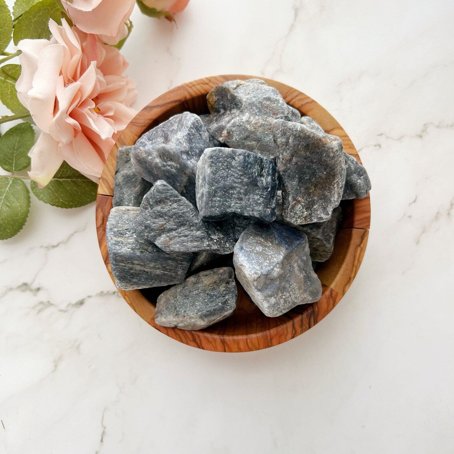 A wooden bowl filled with large chunks of Raw Sapphire Blue Aventurine Stones, known for its third eye chakra properties, sits on a marble surface. Beside the bowl are pink roses with green leaves, adding a soft floral touch to the composition.