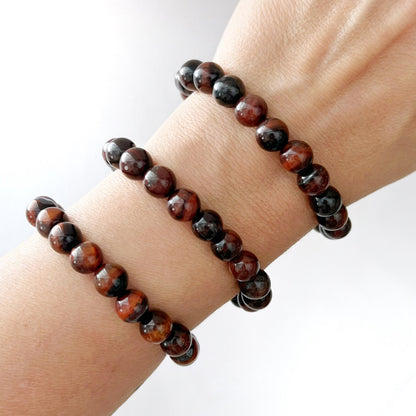 An 8mm Red Tiger Eye Beaded Bracelet featuring alternating dark brown and black polished round stones with Red Tiger Eye accents is laid out on a white surface. In the background, soft pink flowers and more beaded jewelry in a gold dish can be seen.