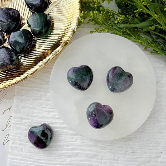 A collection of polished, heart-shaped stones, including 1" Mini Rainbow Fluorite Hearts in shades of green and purple, sits on a round white surface. More stones lie on a textured dish nearby, with sprigs of green foliage in the background. A handwritten note is partially visible on the left.