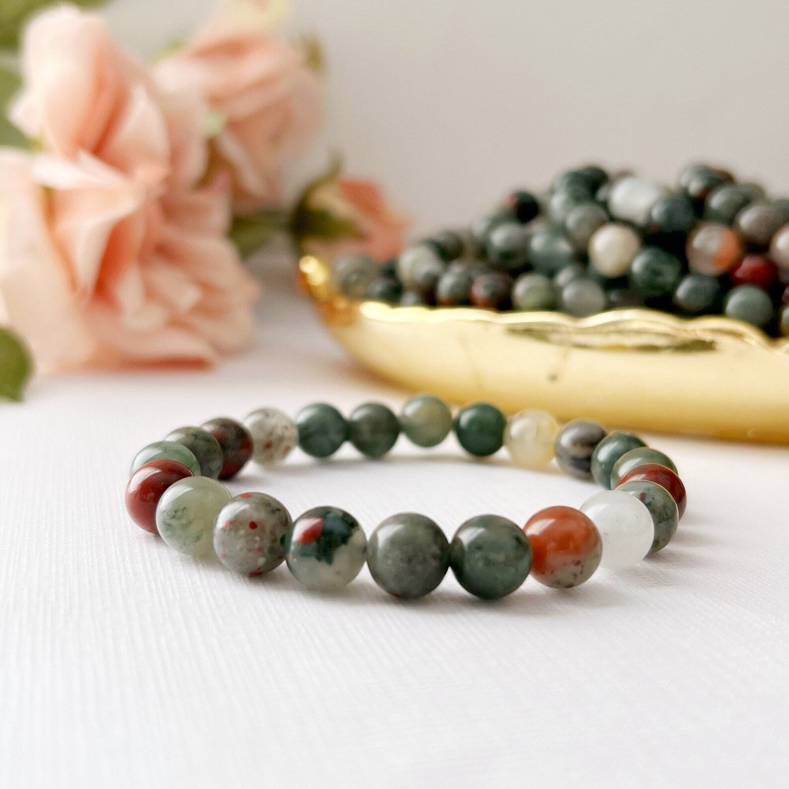 A 8mm Seftonite (African Bloodstone) Beaded Bracelet made of African Bloodstone and multicolored stone beads is displayed on a white surface. Behind the bracelet, there is a golden dish filled with similar beads and soft pink roses, creating an elegant and delicate setting imbued with Usui Reiki energy.