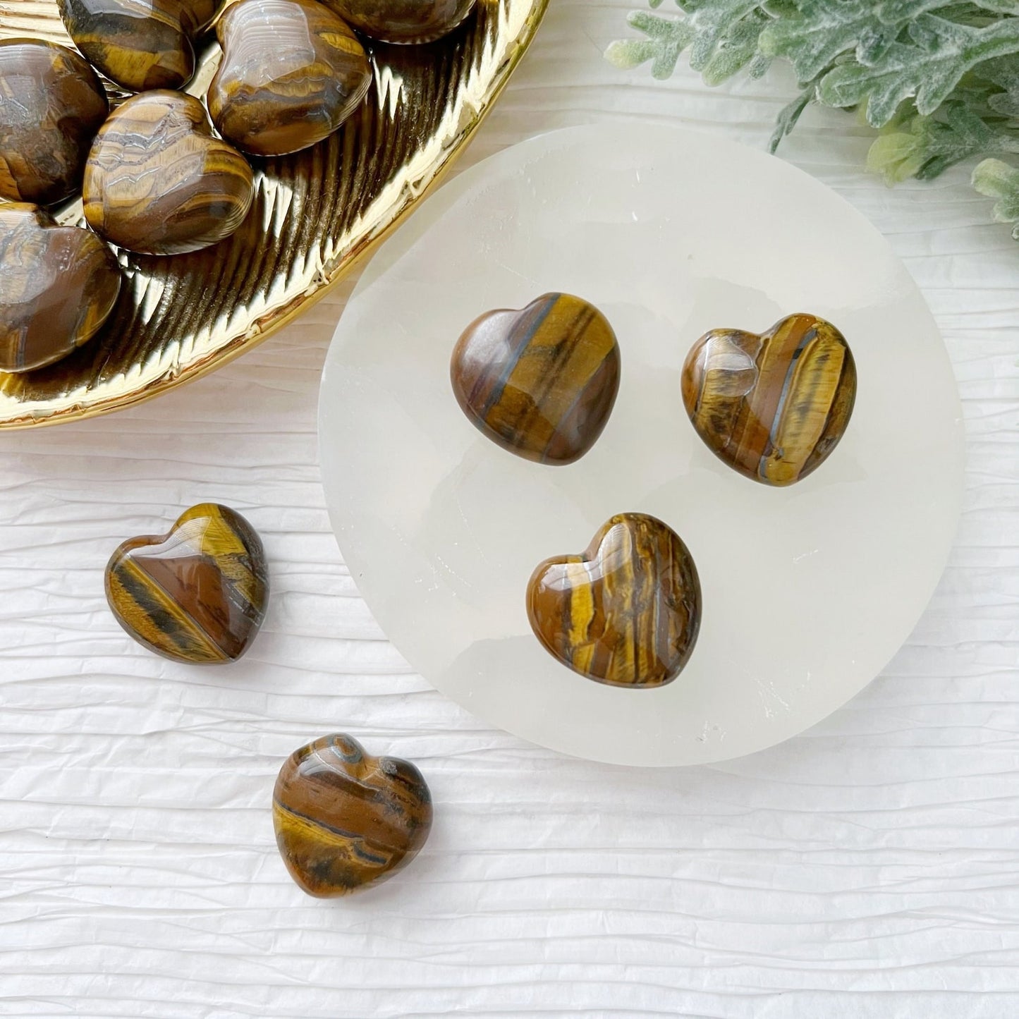 Image of heart-shaped Mini Tiger Iron stones displayed on a white surface. Four puffy hearts are arranged neatly on a round white dish, while several others are placed around and in a nearby gold dish. Green foliage is also visible in the top-right corner, perfect for adding to your crystal grids.