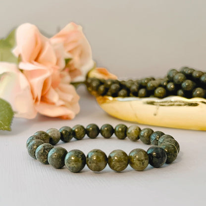 A bracelet made of dark green and gold-speckled round beads, known as an 8mm Epidote Beaded Bracelet, is displayed in the foreground on a light surface. Behind it, a gold dish filled with similar beads and two pink roses are partially visible, adding a touch of elegance to the scene.