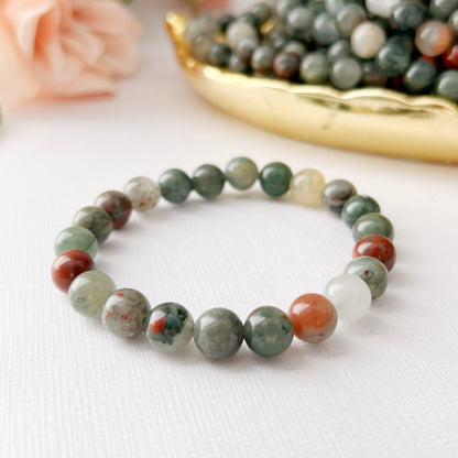 A 8mm Seftonite (African Bloodstone) Beaded Bracelet made of African Bloodstone and multicolored stone beads is displayed on a white surface. Behind the bracelet, there is a golden dish filled with similar beads and soft pink roses, creating an elegant and delicate setting imbued with Usui Reiki energy.