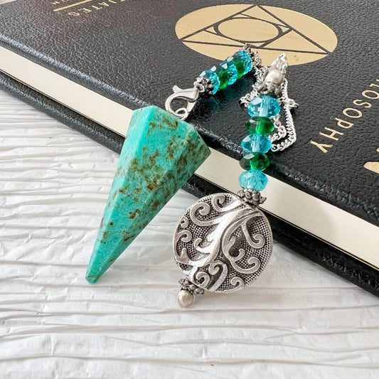 A Chrysocolla Hexagonal Pendulum with Tree of Life Charm is attached to a decorative chain with green and clear beads. Next to it is a silver pendant featuring a Tree of Life charm with intricate swirling patterns. Both items are placed on a closed book with a geometric symbol on its cover, perfect for use as divination tools.