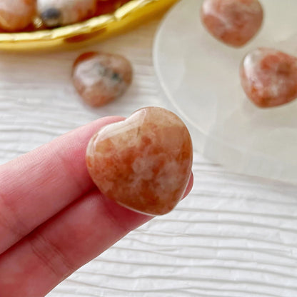 Heart-shaped, peach-colored crystals are displayed on a white textured surface. Four 1" Mini Sunstone Heart crystals are arranged on a round white plate, with a golden dish holding several more 1" Mini Sunstone Hearts in the background. Green foliage partially visible at the top right adds a natural touch.