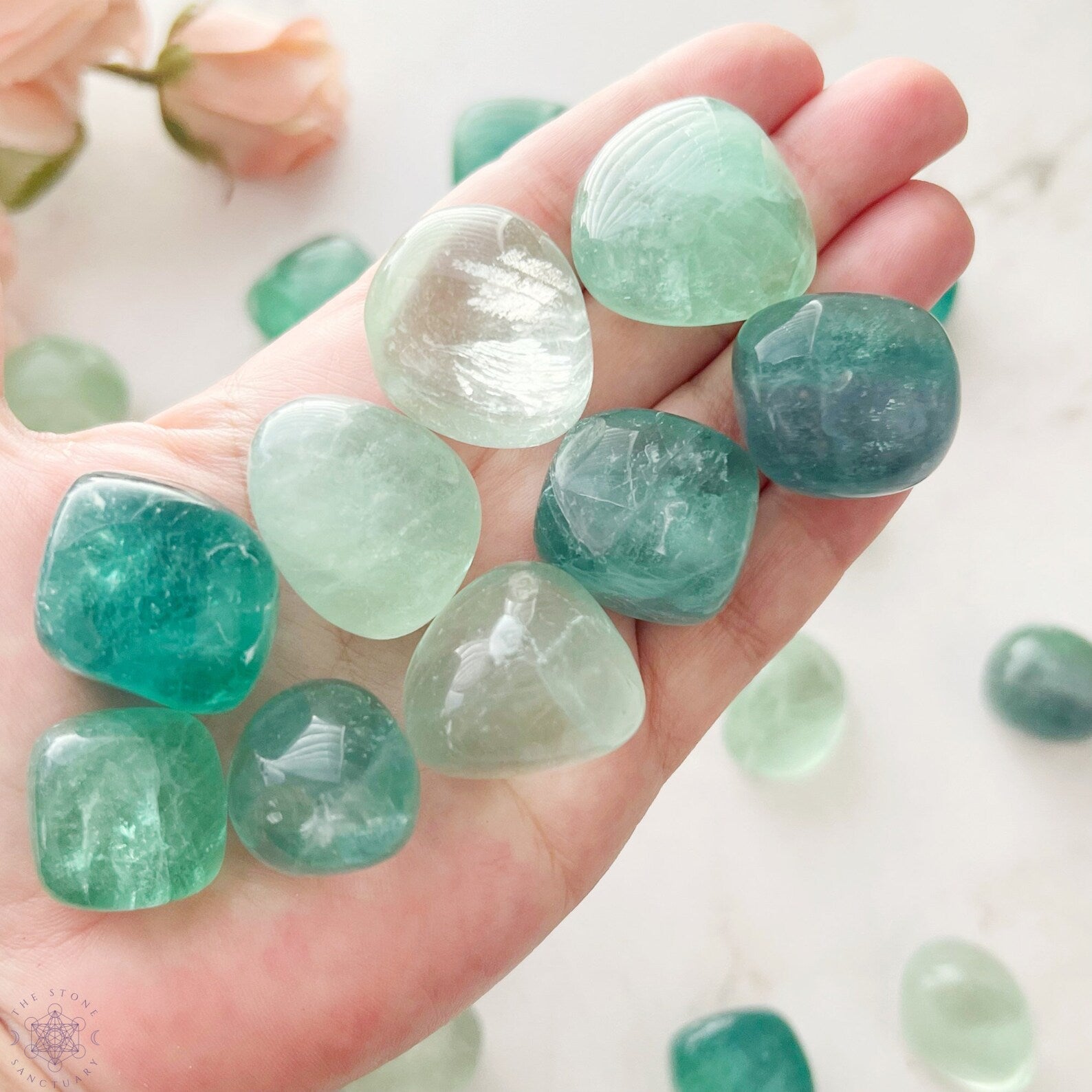 A wooden bowl is filled with various polished translucent green stones, including stunning Green Fluorite Tumbled Stones. Some have faint white or darker green swirls. The stones are smooth and glossy, reflecting light with a natural luster.