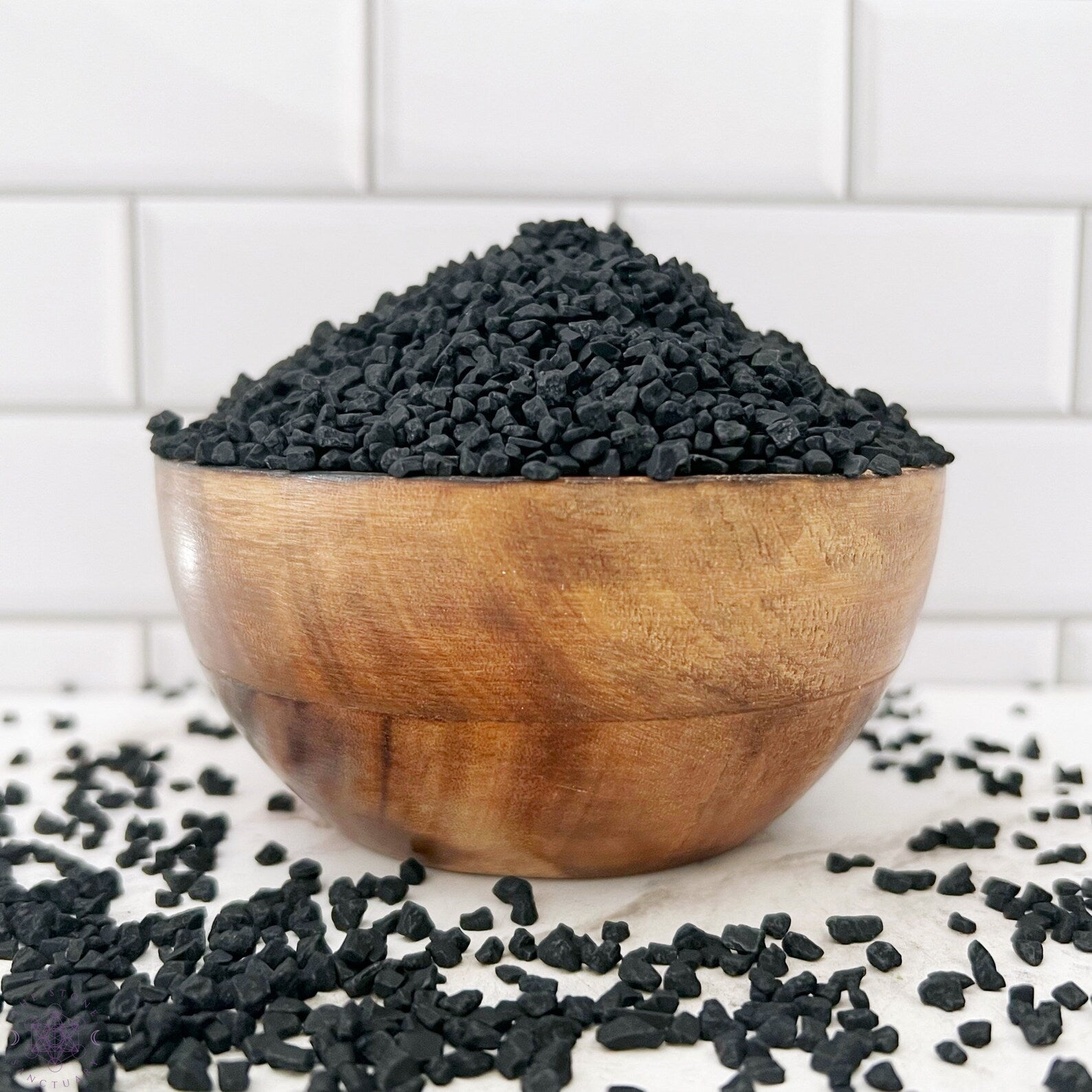 A small sealed package labeled "Black Lava Sea Salt" is placed in front of a glass bowl filled with black salt and a white mortar and pestle. The background features white tiled walls, hinting at the enchanting allure of using Black Salt for Spells with its touch of activated charcoal.