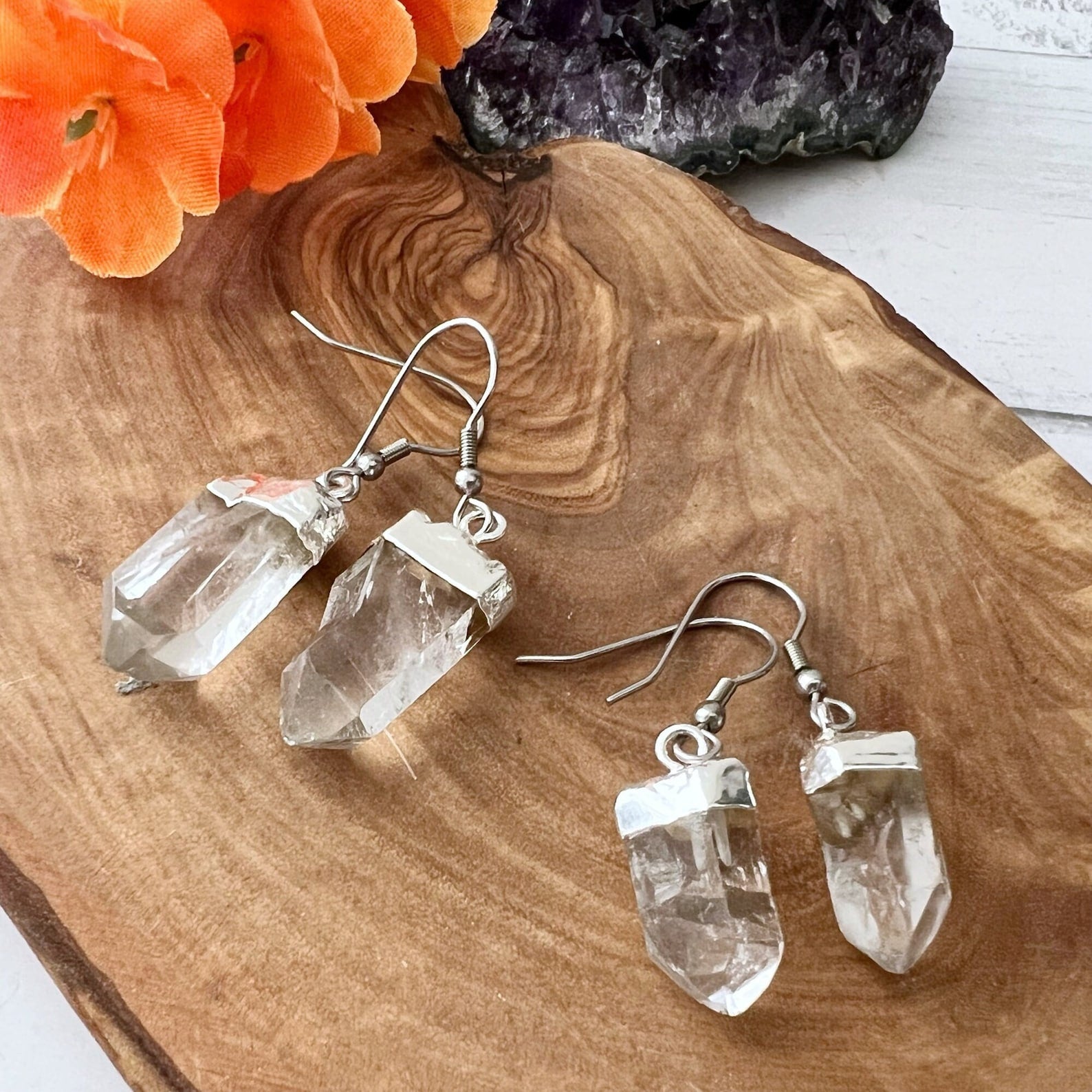 A pair of natural crystal earrings with silver hooks are displayed on a wooden surface. An orange flower and a piece of amethyst crystal are visible in the background, adding contrast and color to the scene. These unique Raw Clear Quartz Point Dangle Earrings make for stunning jewelry pieces.