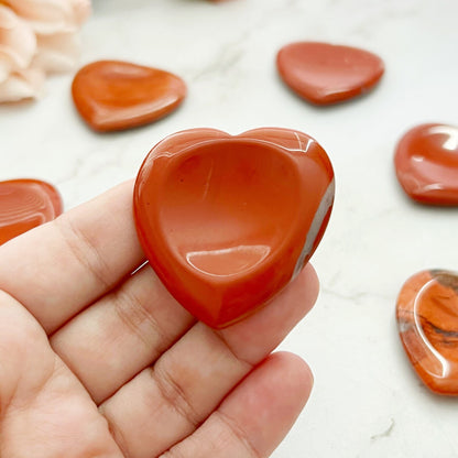 Five polished red heart-shaped stones, including Red Jasper Heart Shaped Worry Stones, are displayed on a wooden tray, surrounded by a white marble surface. The wooden tray rests below a light pink rose with green leaves. Some stones are on the tray, while others are scattered across the marble surface.