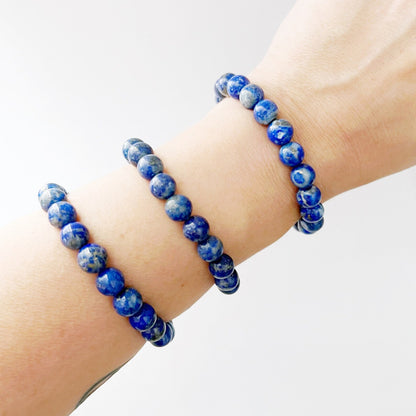 An 8mm Lapis Lazuli Beaded Bracelet made of rounded beads is displayed on a white surface. Behind it, there are two light pink roses and a gold dish filled with more blue beads, all slightly out of focus.