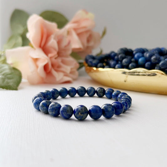 An 8mm Lapis Lazuli Beaded Bracelet made of rounded beads is displayed on a white surface. Behind it, there are two light pink roses and a gold dish filled with more blue beads, all slightly out of focus.