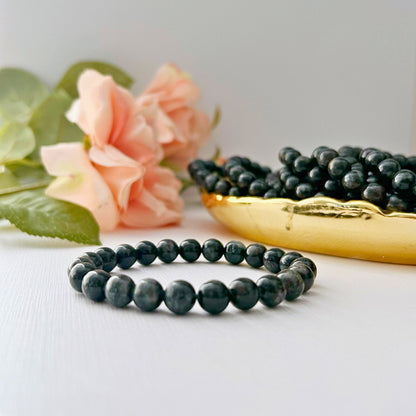 An 8mm Astrophyllite Bead Bracelet, crafted from natural gemstone beads, is prominently displayed in the foreground against a white background. Behind it, a golden dish filled with more black beads is partially visible. To the left, pink roses and green leaves add a touch of natural elegance to the scene.