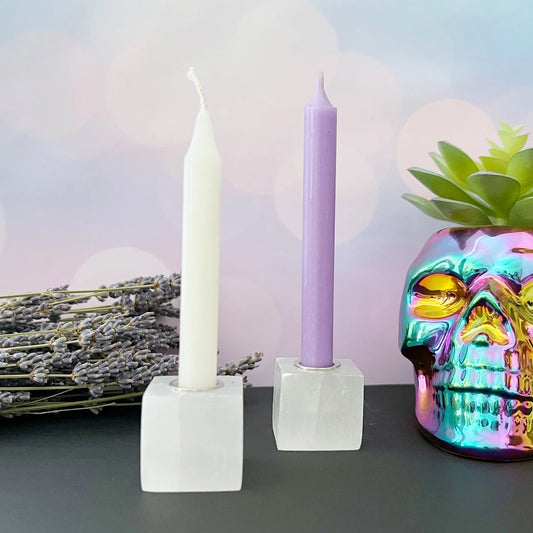 A pastel-themed arrangement featuring using a 1" Selenite Cube Mini Candle Holder for Ritual Candles, two Chime Spell Candles—one in white and one in lavender—a bundle of lavender flowers, and a prismatic skull-shaped vase containing a small green succulent. This setup provides the perfect ambiance for meditation or spiritual work amidst the soft bokeh background with pale colors.