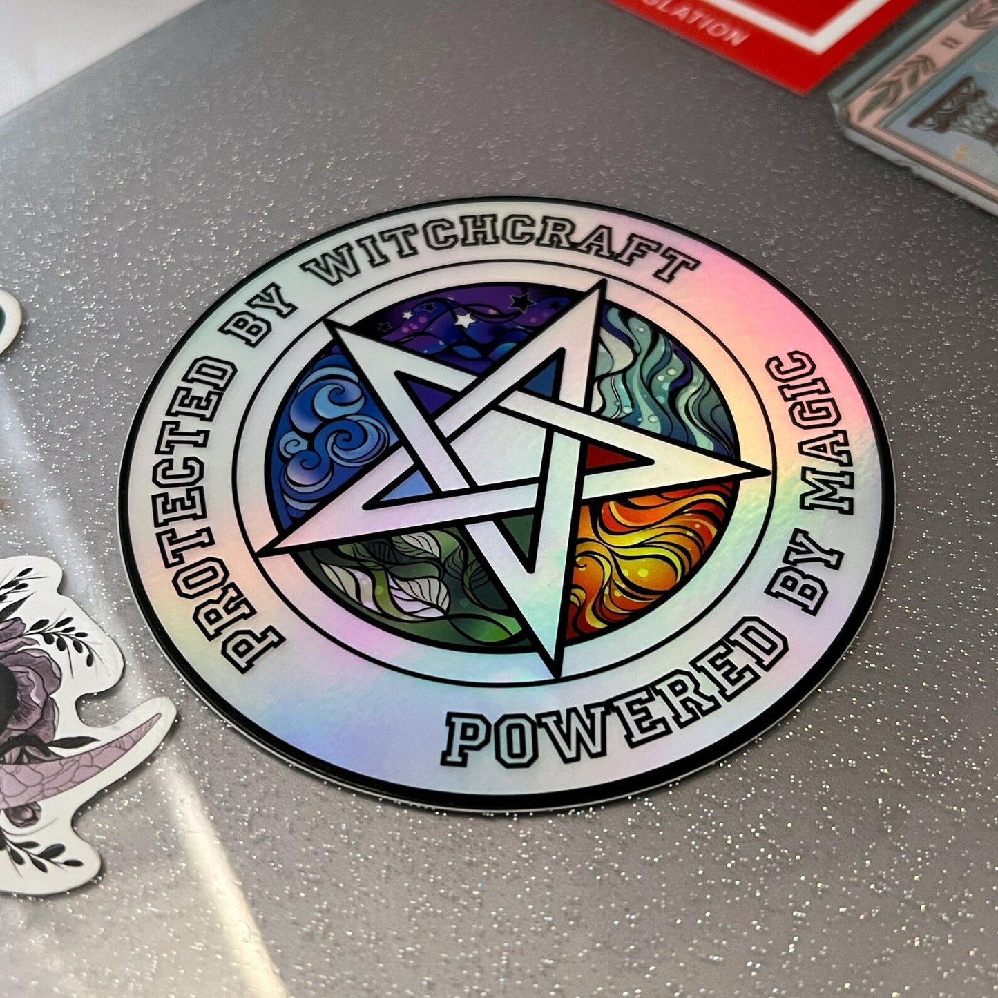 A hand holds a shiny, iridescent 4" Holographic Protected by Witchcraft Sticker with a colorful pentagram in the center. The text on the holographic vinyl sticker reads, "Protected by Witchcraft, Powered by Magic." The background features a soft bokeh effect with pastel colors.