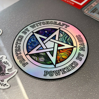 A hand holds a shiny, iridescent 4" Holographic Protected by Witchcraft Sticker with a colorful pentagram in the center. The text on the holographic vinyl sticker reads, "Protected by Witchcraft, Powered by Magic." The background features a soft bokeh effect with pastel colors.