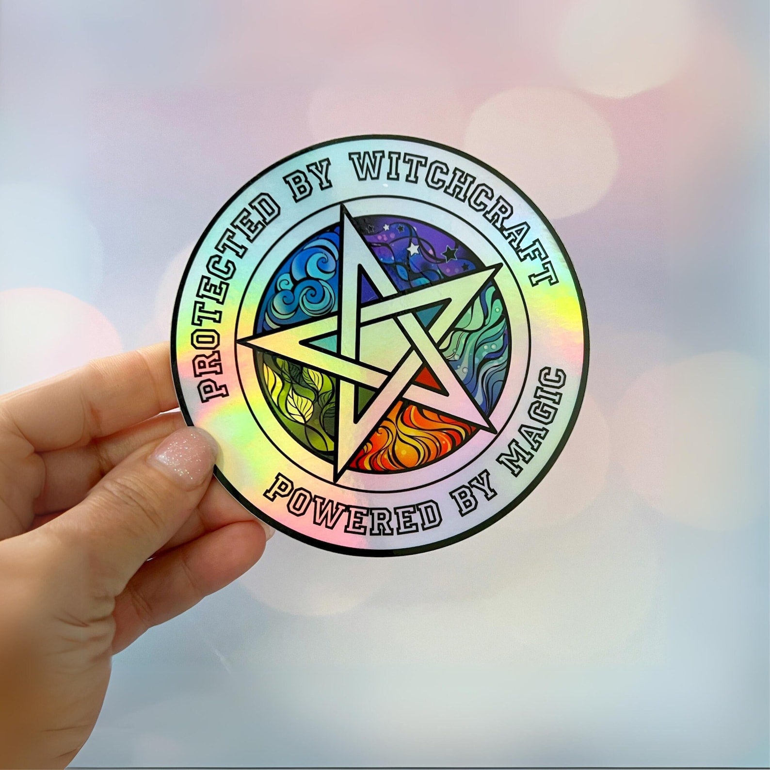 A hand holds a shiny, iridescent 4" Holographic Protected by Witchcraft Sticker with a colorful pentagram in the center. The text on the holographic vinyl sticker reads, "Protected by Witchcraft, Powered by Magic." The background features a soft bokeh effect with pastel colors.