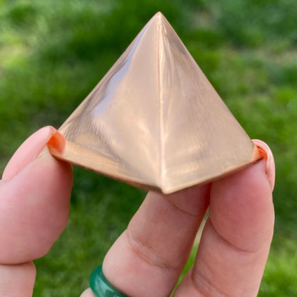 A hand with pinkish nails and a green ring holds a small, metallic 1" Copper Pyramid with a gleaming surface, perfect for meditation and energy work. The background is blurred and grassy.