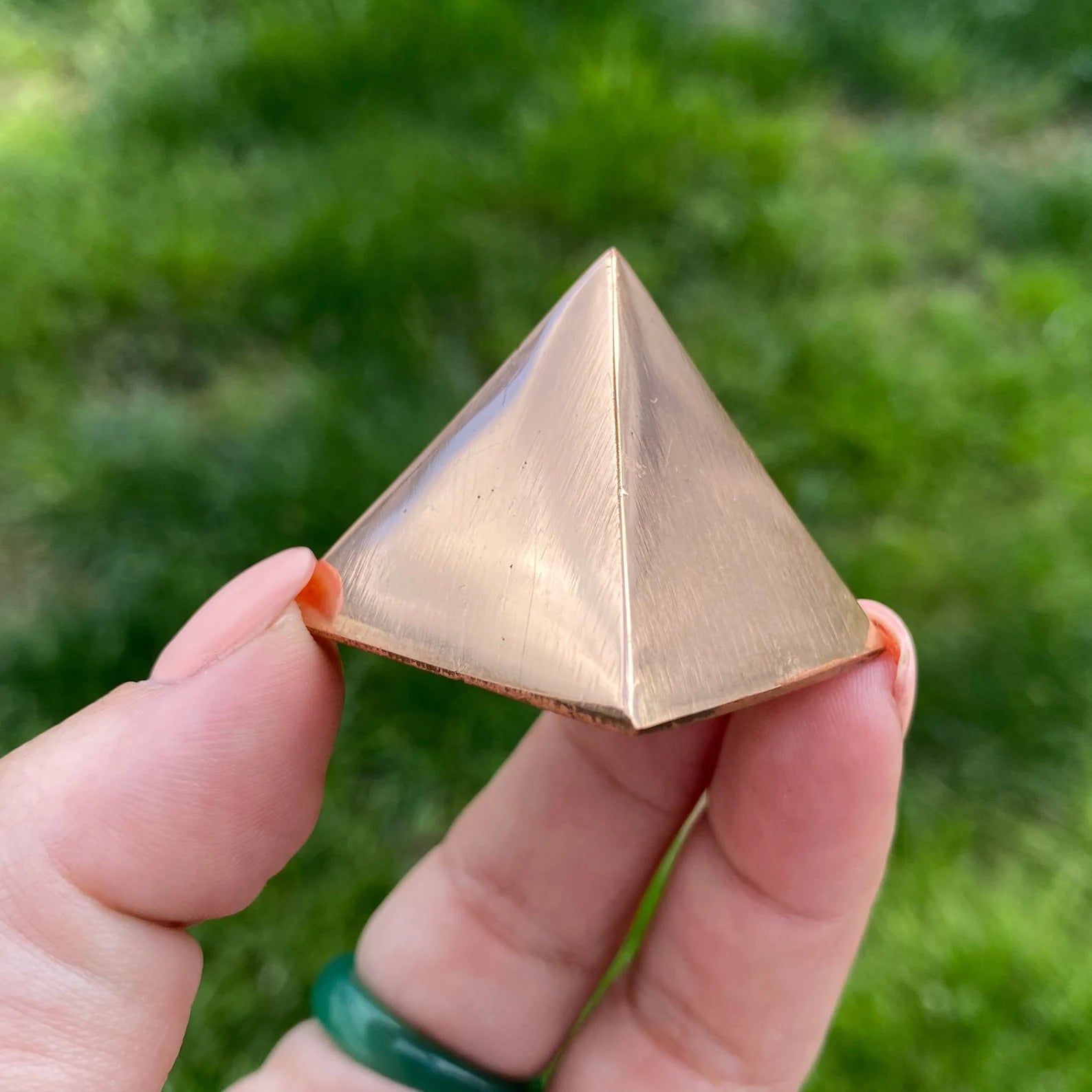 A hand with pinkish nails and a green ring holds a small, metallic 1" Copper Pyramid with a gleaming surface, perfect for meditation and energy work. The background is blurred and grassy.