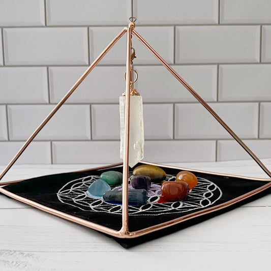 A 6" Copper Pyramid Energizer w/ Quartz Point and Mat encloses a collection of colorful crystals. The crystals rest on a black cloth adorned with a white geometric pattern, akin to a chakra mat, placed against a white tiled background.