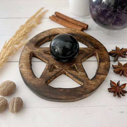 A black crystal sphere sits on a Wooden Pentacle Sphere Stand. Surrounding the stand are star anise, nutmeg, a purple crystal ball holder, dried wheat stalks, and cinnamon sticks, all arranged on a light wooden surface.