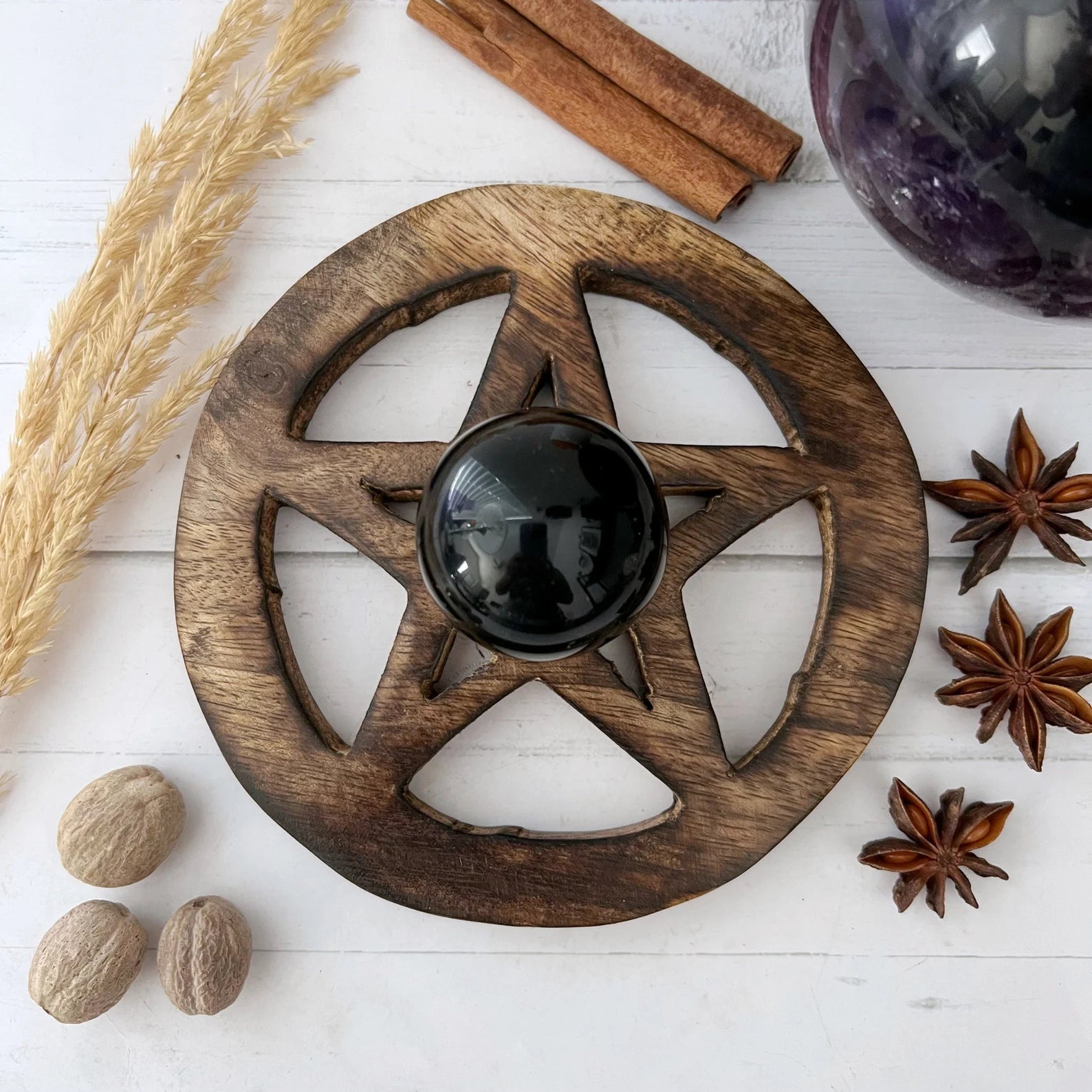 A black crystal sphere sits on a Wooden Pentacle Sphere Stand. Surrounding the stand are star anise, nutmeg, a purple crystal ball holder, dried wheat stalks, and cinnamon sticks, all arranged on a light wooden surface.