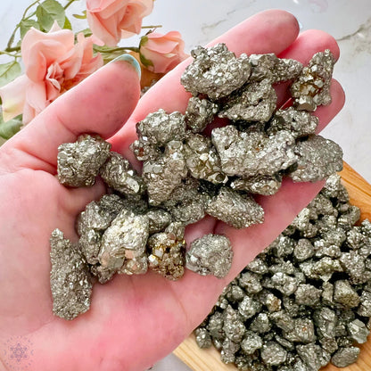 A wooden tray filled with shiny, metallic 12-18mm Pyrite Crystal Chips is placed on a marble surface. Some raw and unpolished stones are scattered beside the tray. Pink roses are positioned at the top left corner, partially in the frame.